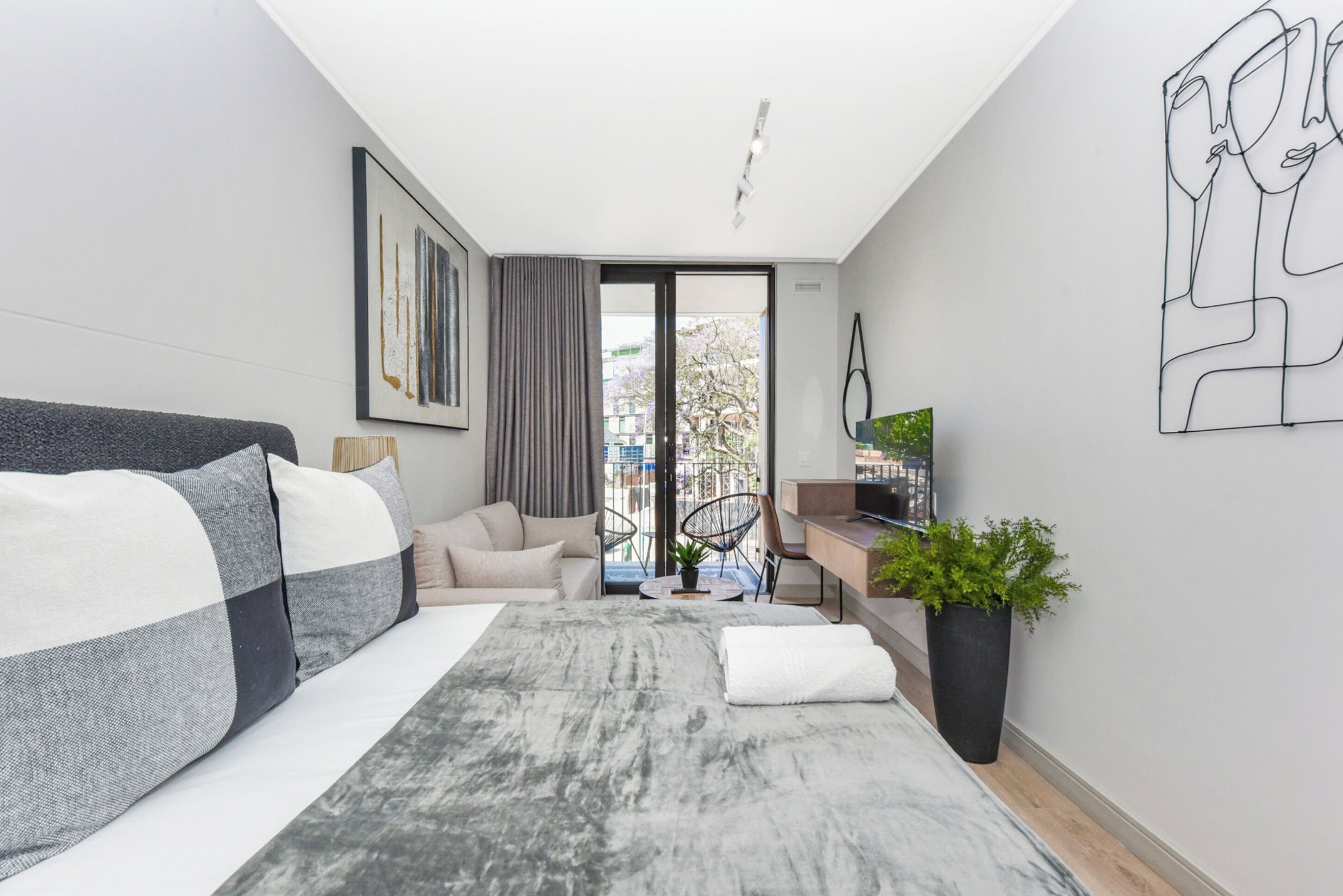 Una habitación de hotel moderna con una cama grande, un escritorio con computadora, un televisor y un balcón con vista a la ciudad.