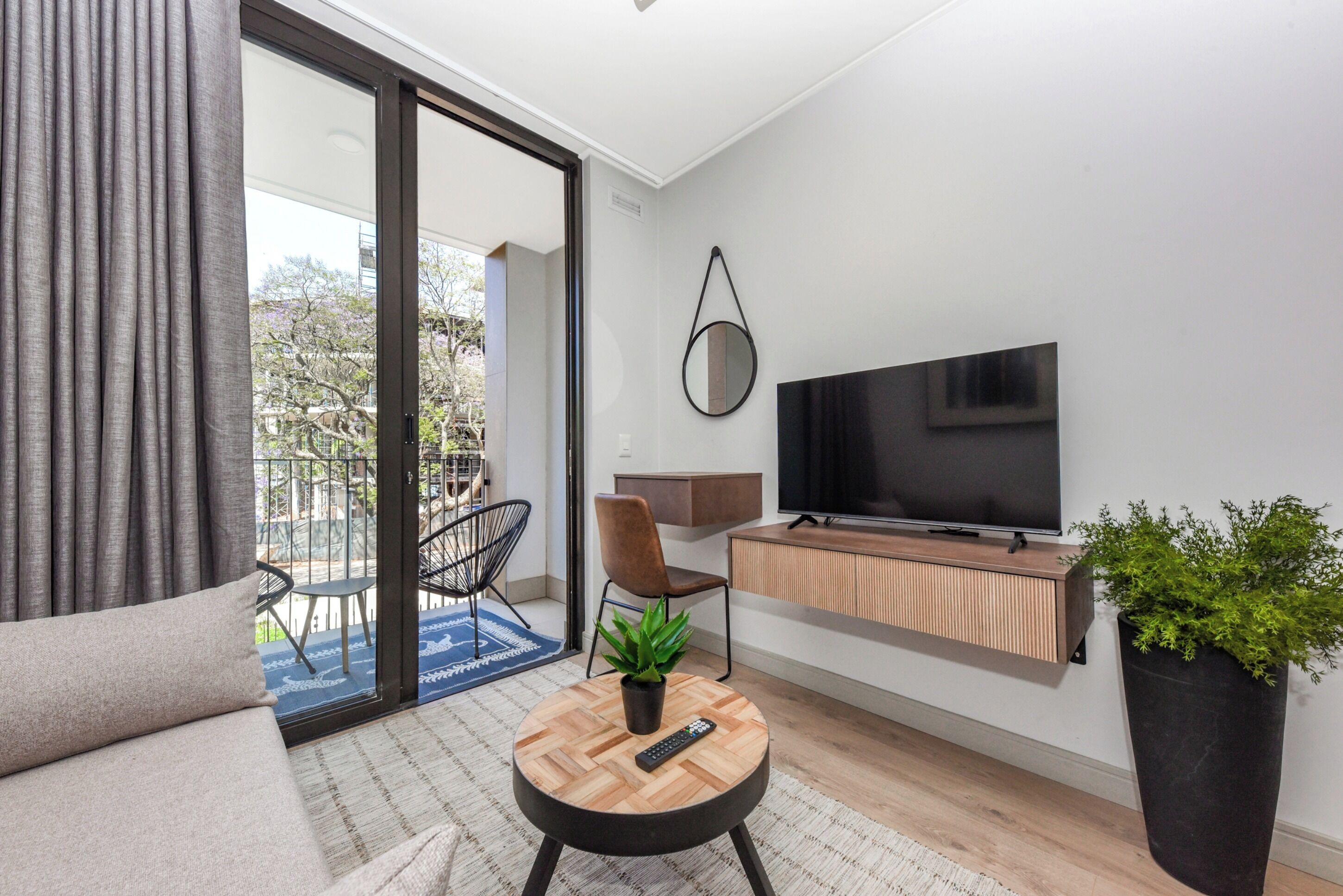 Una sala de estar moderna con un sofá, una mesa redonda de centro con un control remoto, un televisor sobre un mueble de madera, una planta en maceta y un puerta corrediza de vidrio que da a un balcón.