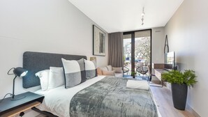 A modern hotel room with a large bed, a desk with a lamp, a potted plant, and a view of the outdoors.