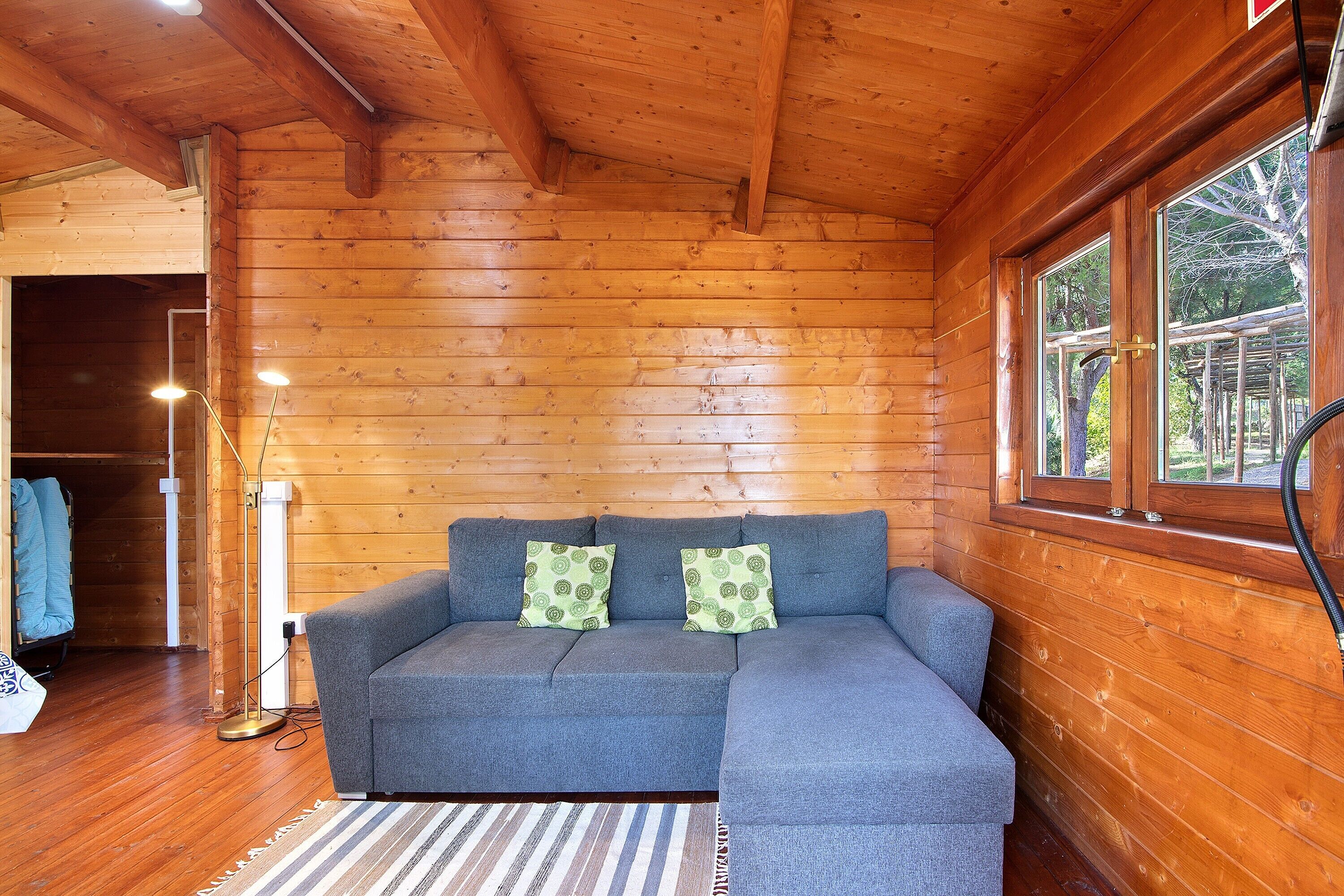A wooden cabin interior with a gray sectional sofa, two patterned pillows, a standing floor lamp, and a window with a view of trees.
