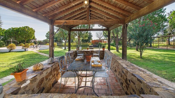 An outdoor covered patio with a stone wall, metal table and chairs, and a hanging light fixture. - Casa de Campo Vale do Asno – Bungalow '2' with Shared Pool, Wi-Fi & Air Conditioning (Castro Marim)