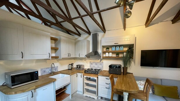 A compact kitchen with white cabinets, a microwave, a stove, and a sink. There is a wooden dining table with chairs and a sofa with a yellow pillow.