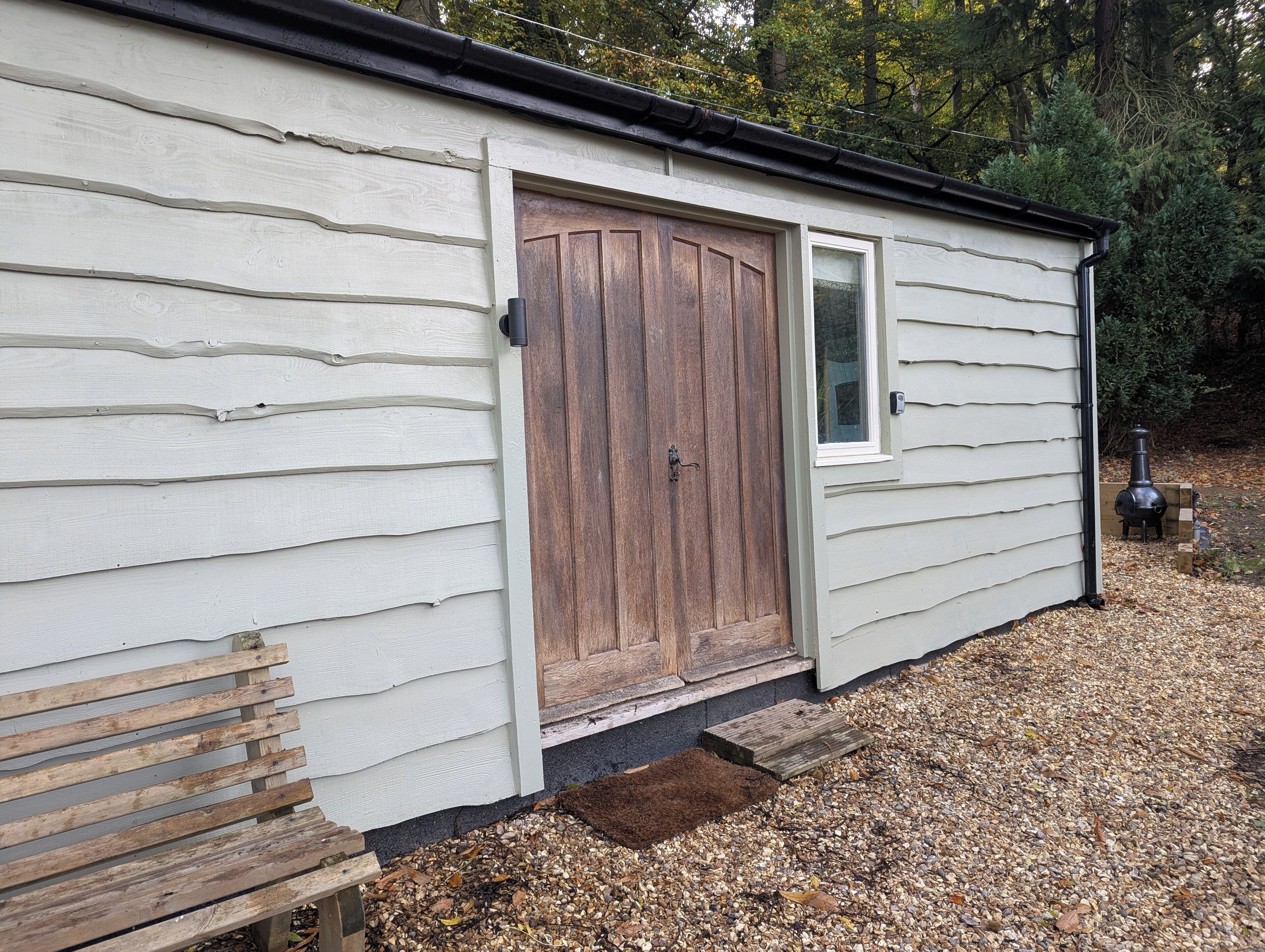 A small wooden shed with a single door and a window, surrounded by gravel and trees.