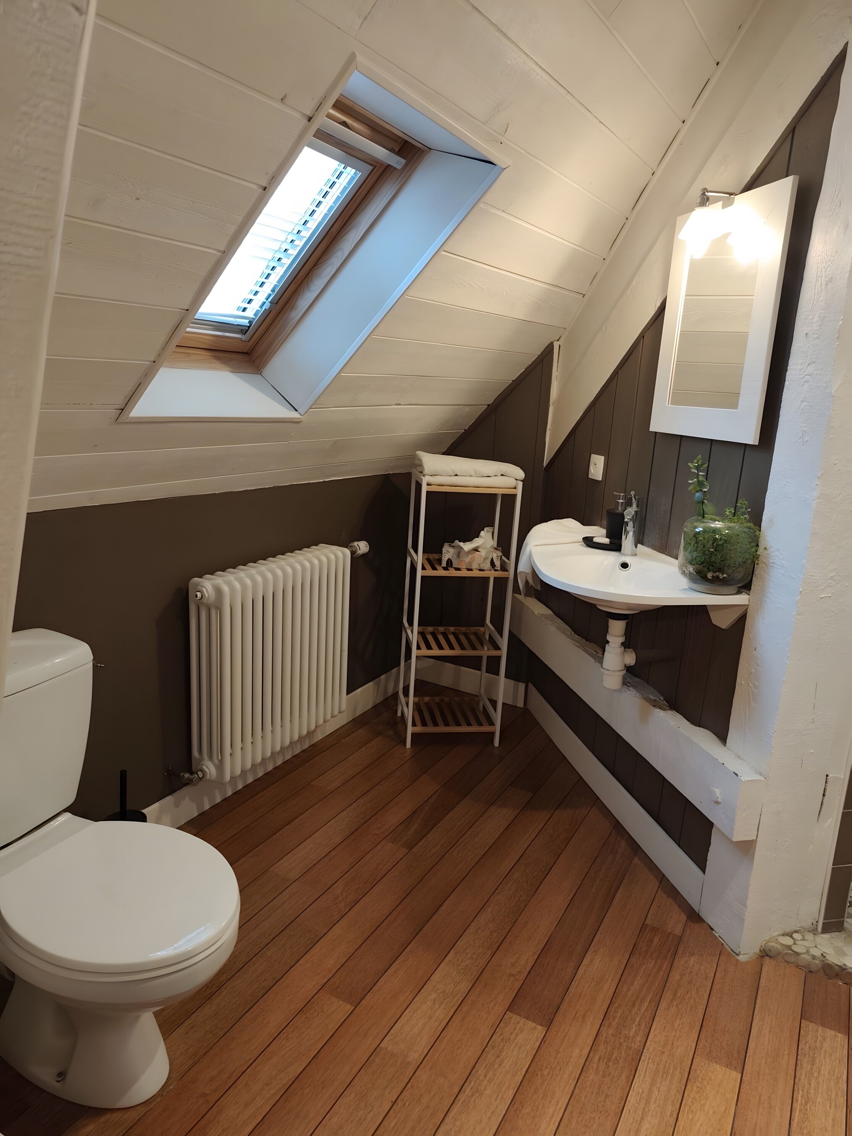 A small, sloped-roofed bathroom with a toilet, a white sink, a mirror, and a radiator.