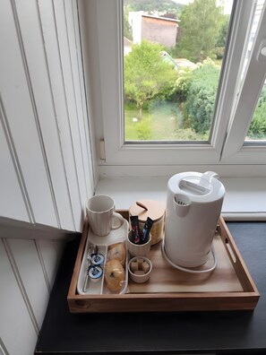 Una bandeja con una cafetera, taza y recipientes de bocadillos junto a una ventana con vista a follaje.
