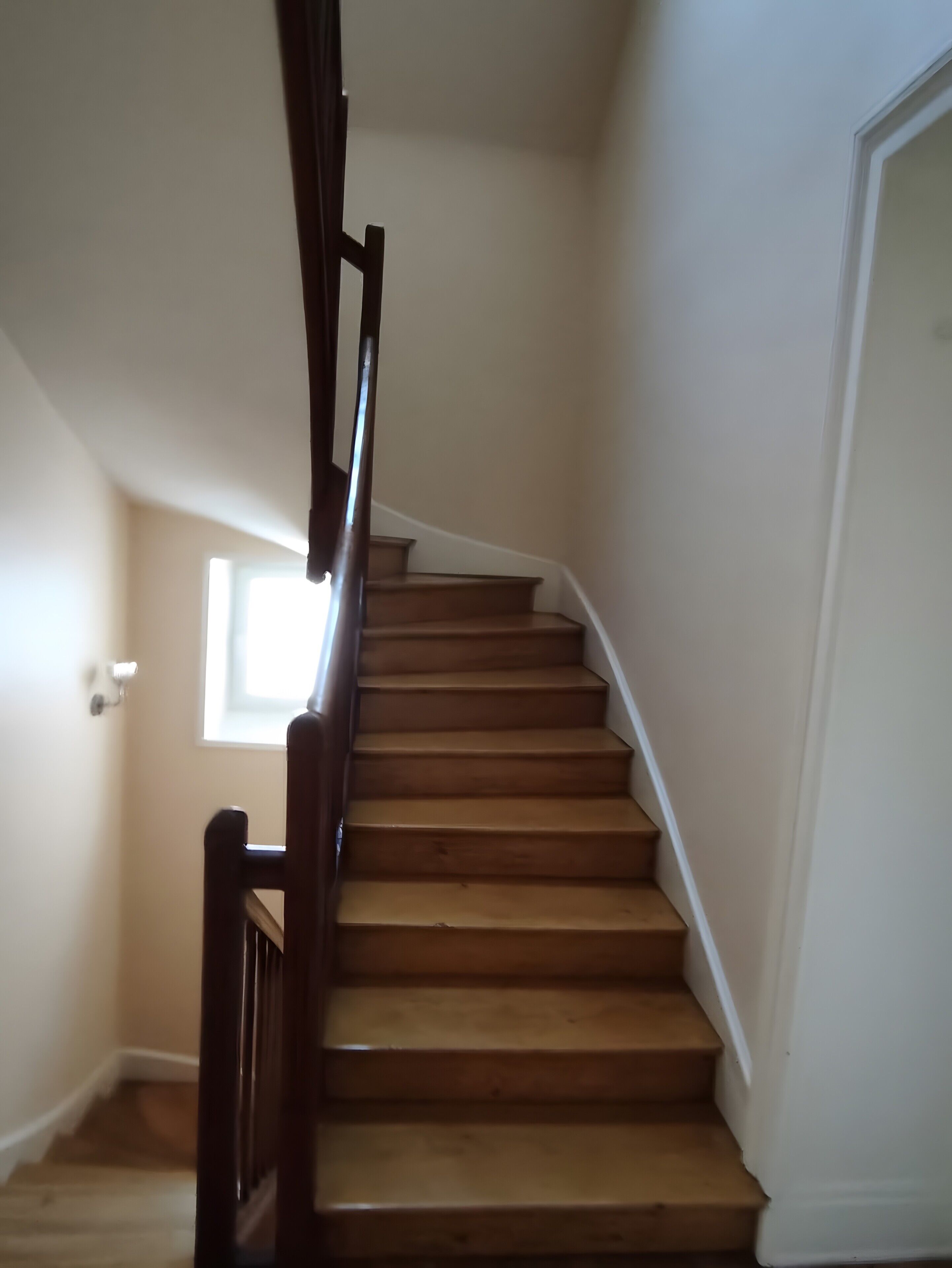 Una escalera de madera con barandilla, que lleva a un rellano con una puerta blanca y un ventanal pequeño.