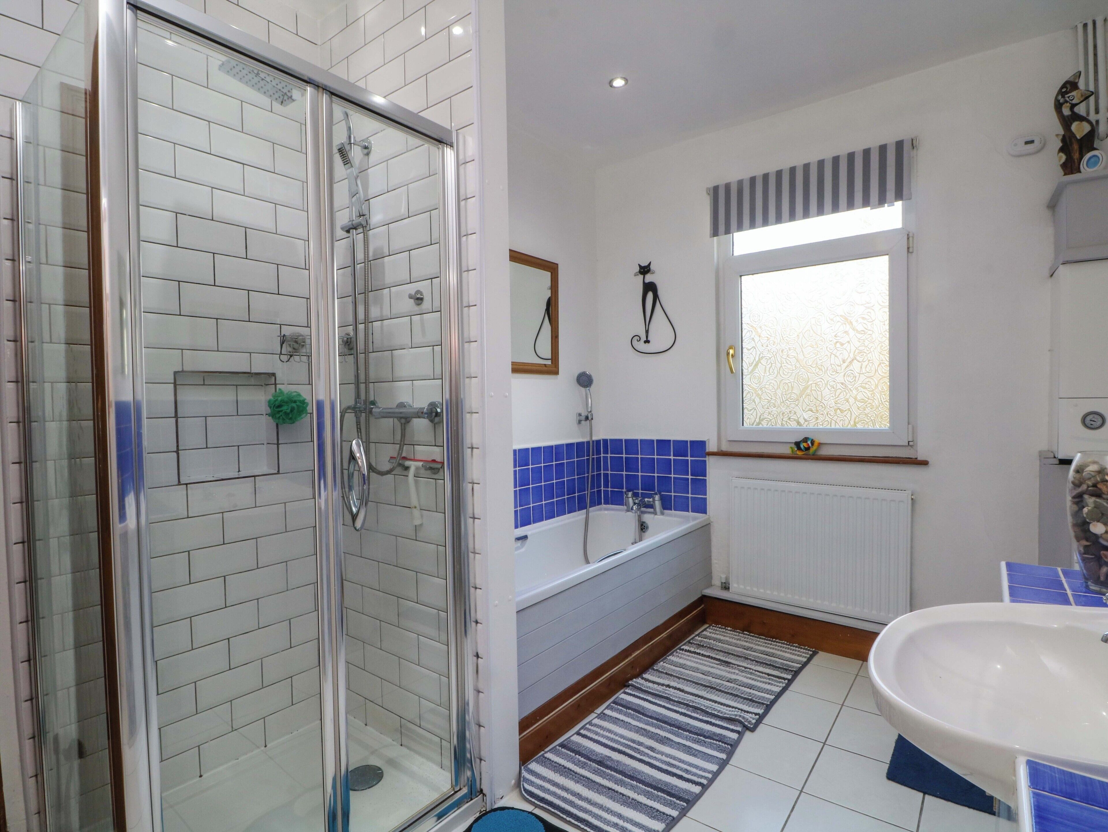 A bathroom with a glass shower enclosure, a white sink, a blue-tiled bathtub, and a window with a striped curtain.
