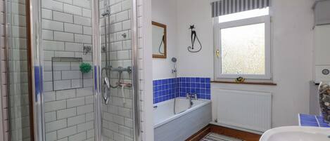 A bathroom with a glass shower enclosure, a white sink, a blue-tiled bathtub, and a window with a striped curtain.