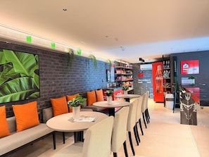 Un café moderne avec un mur en briques, des plantes vertes, des coussins orange et un réfrigérateur Coca-Cola.