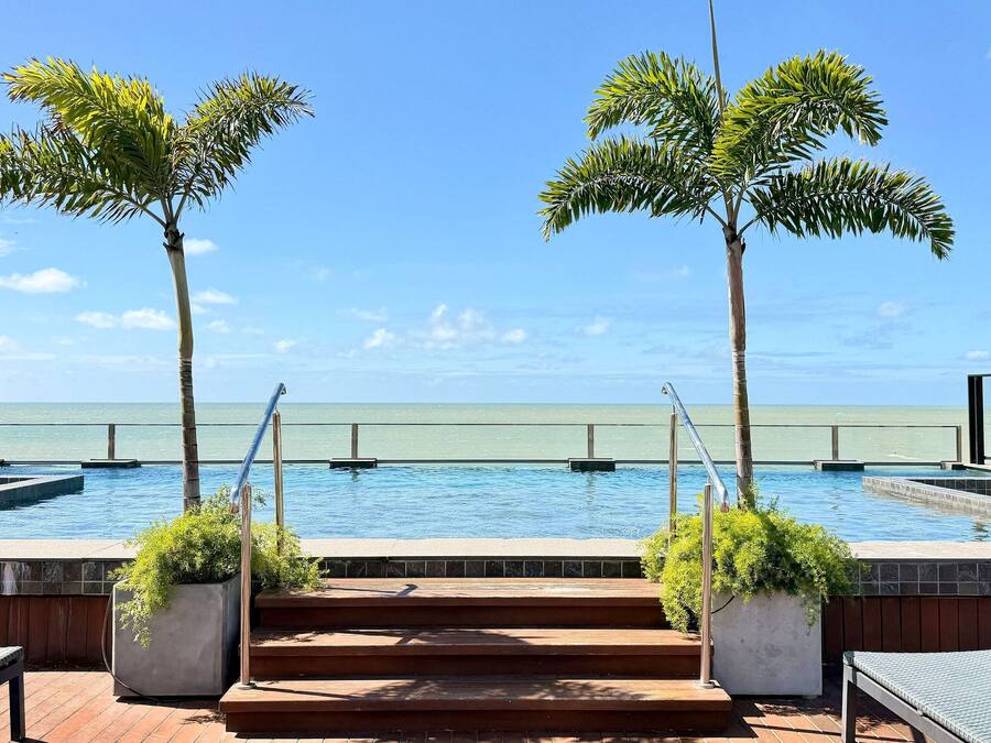 A pool area with steps leading to the water, surrounded by palm trees and a view of the ocean.