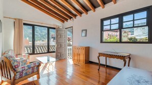 Um quarto espaçoso com piso de madeira, uma mesa de vidro, um sofá com estofamento floral e vista para uma paisagem montanhosa através de amplas janelas.