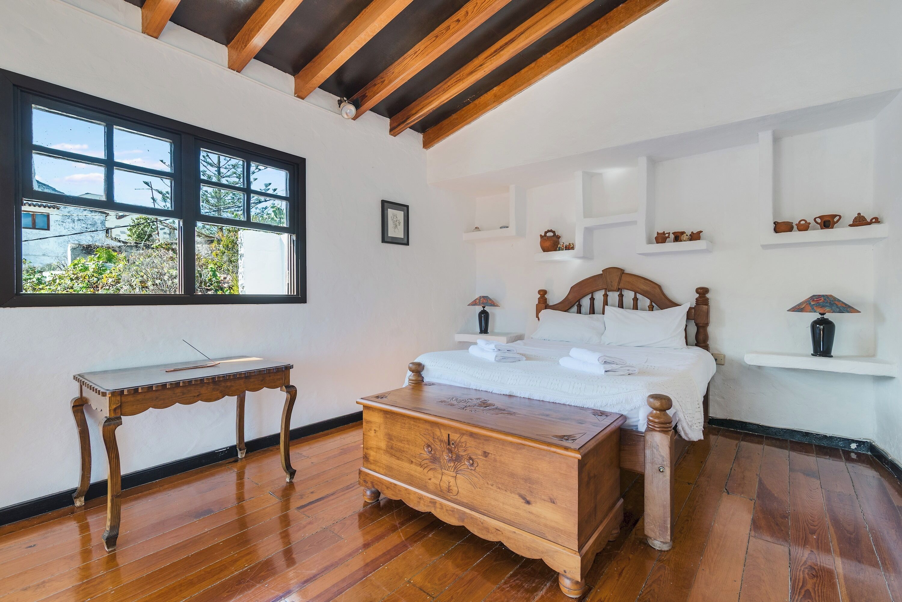 A bedroom with a wooden bed, a nightstand, a dresser, and a window with a view of greenery.