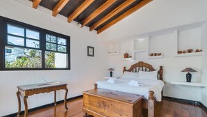 Quarto com cama de madeira, mesa de cabeceira, cômoda e janela com vista para vegetação.