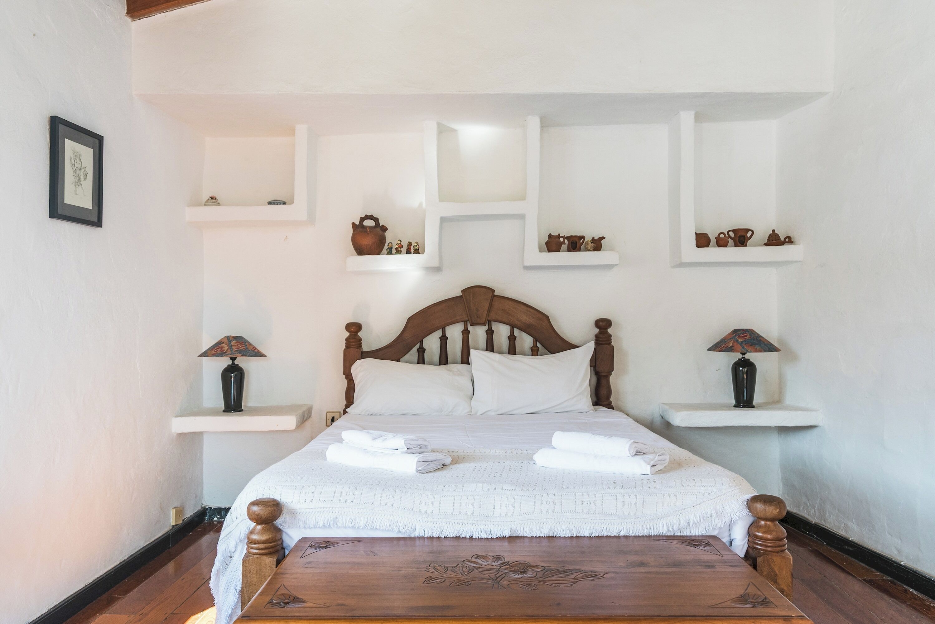 A neatly made bed with white linens and a wooden headboard. There are two bedside tables with lamps and shelves above the bed displaying various items.