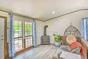A room with a stone wall, a wood-burning stove, a hanging chair, and a view through large windows. - Walk to Water! Blue Lake House w/ Fenced Yard (Quinlan)