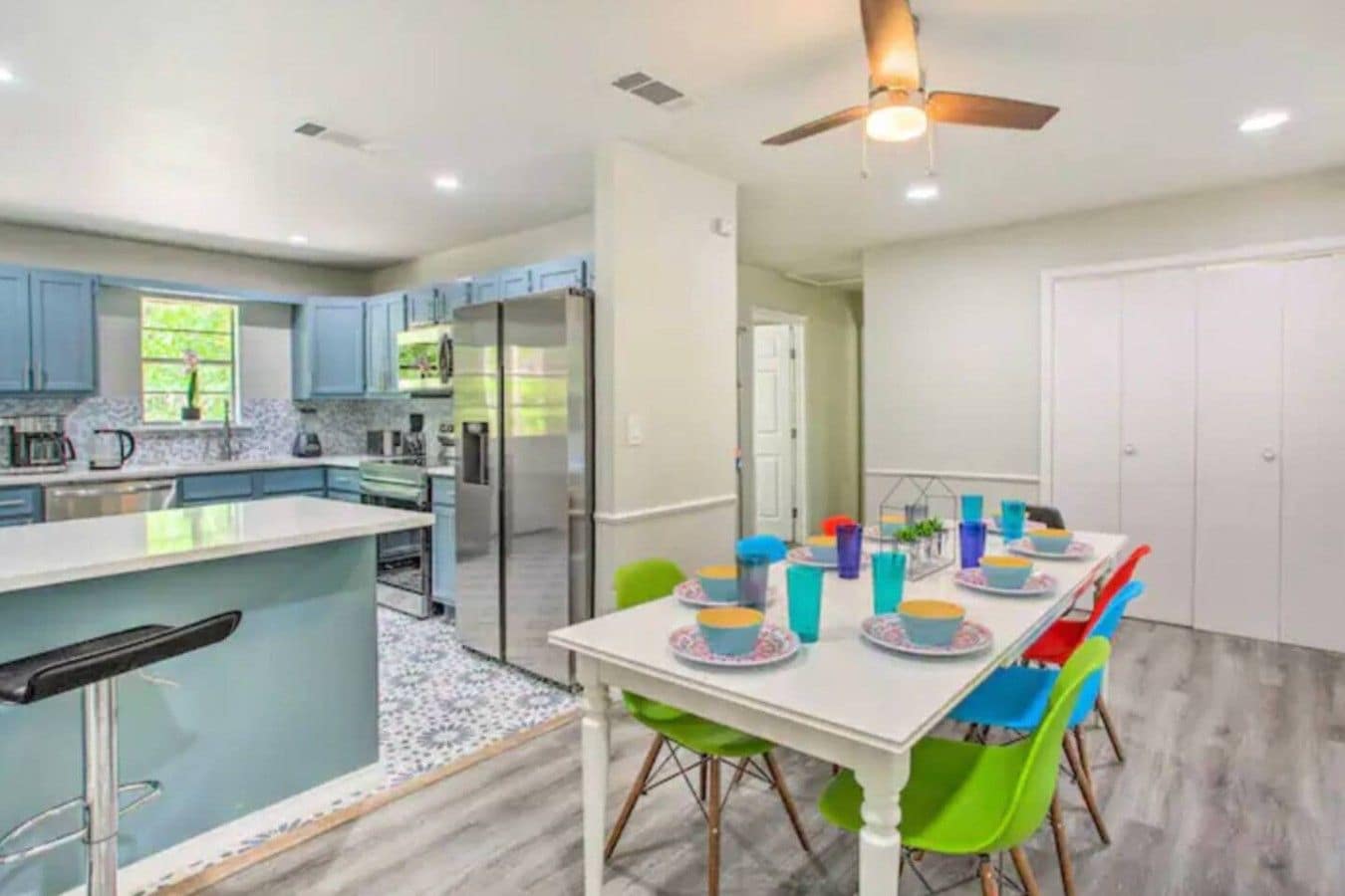 A modern kitchen with a dining area, featuring a central island, a ceiling fan, and a dining table set with colorful chairs and plates.