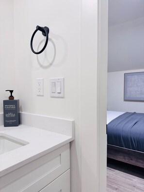 A bathroom with a white sink, a bottle of whiskey leather shampoo, a black towel ring, and a bed with a blue blanket in the adjacent room. - Deluxe Queen Room with Ensuite Bathroom Vancouver West (Vancouver)