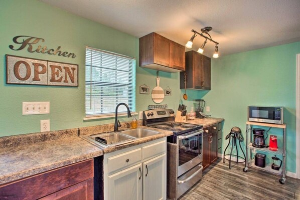 A kitchen with a sink, stove, oven, and cabinets. There is a window with blinds, a ceiling light fixture, and a sign on the wall.