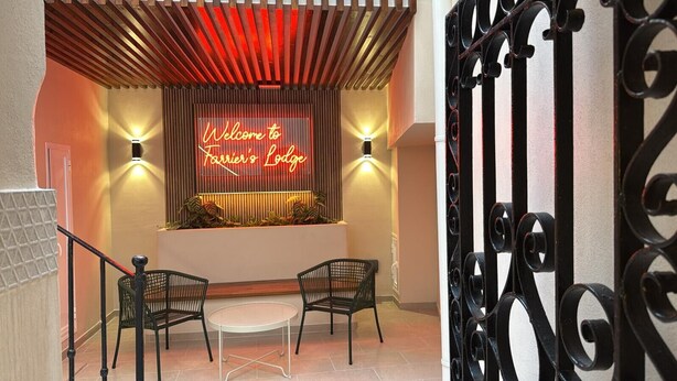 Un área de recepción con un letrero de neón que dice "Welcome to Farrier's Lodge", dos sillones de mimbre, una pequeña mesa redonda y una puerta metálica decorativa.