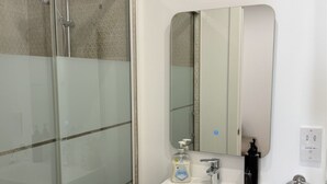 Un baño con cabina de ducha de vidrio, un lavabo blanco con un espejo encima y un perchero con una toalla.