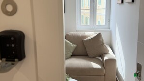 Una habitación con un sofá color beige, una mesita redonda, una puerta con un número y una ventana con vistas a edificios.