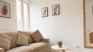 Un salón con un sofá color beige, una mesita redonda con una planta y dos cuadros en la pared.