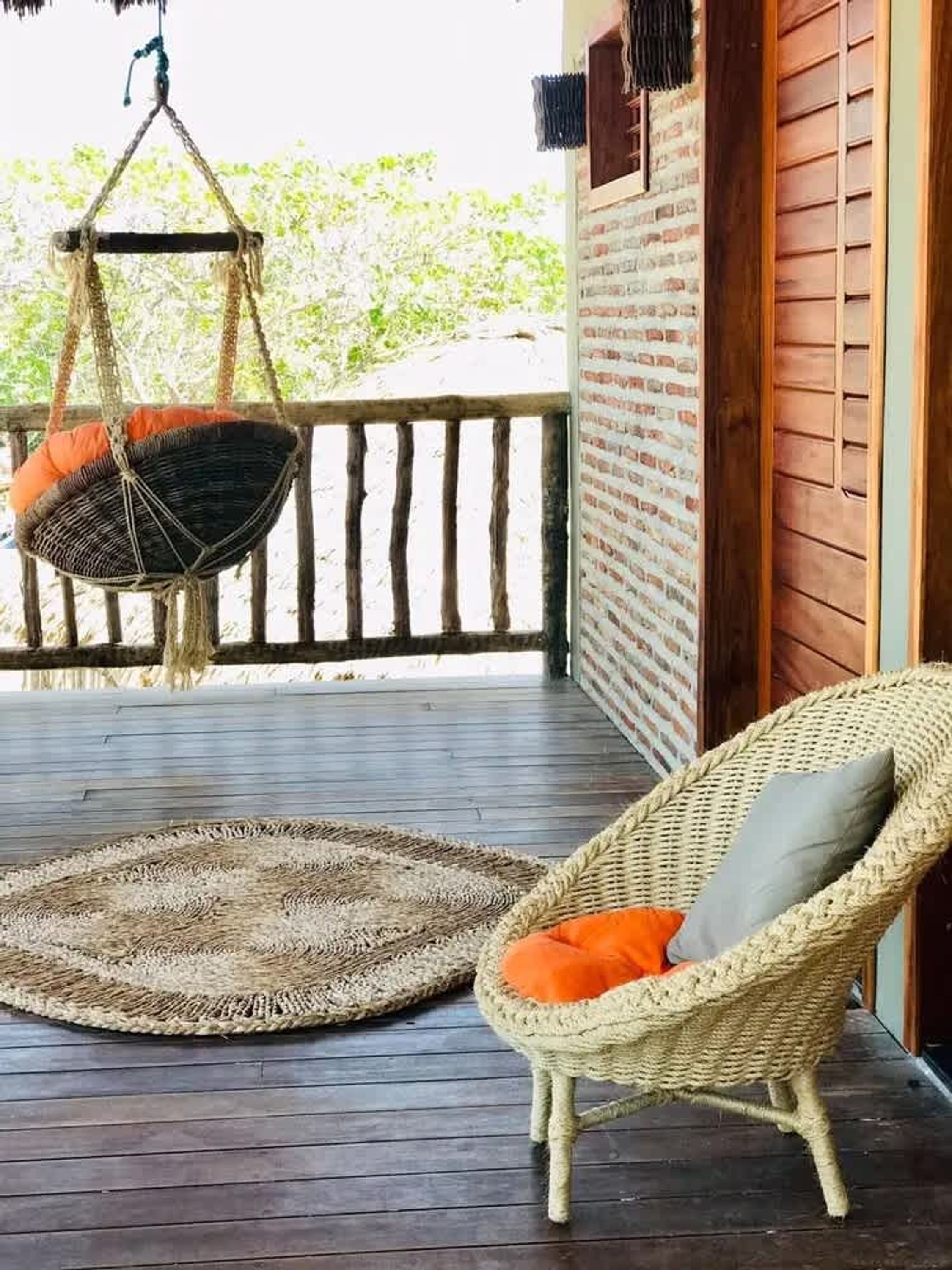A wicker chair with an orange cushion on a wooden deck, a hanging chair with an orange cushion, and a woven rug.
