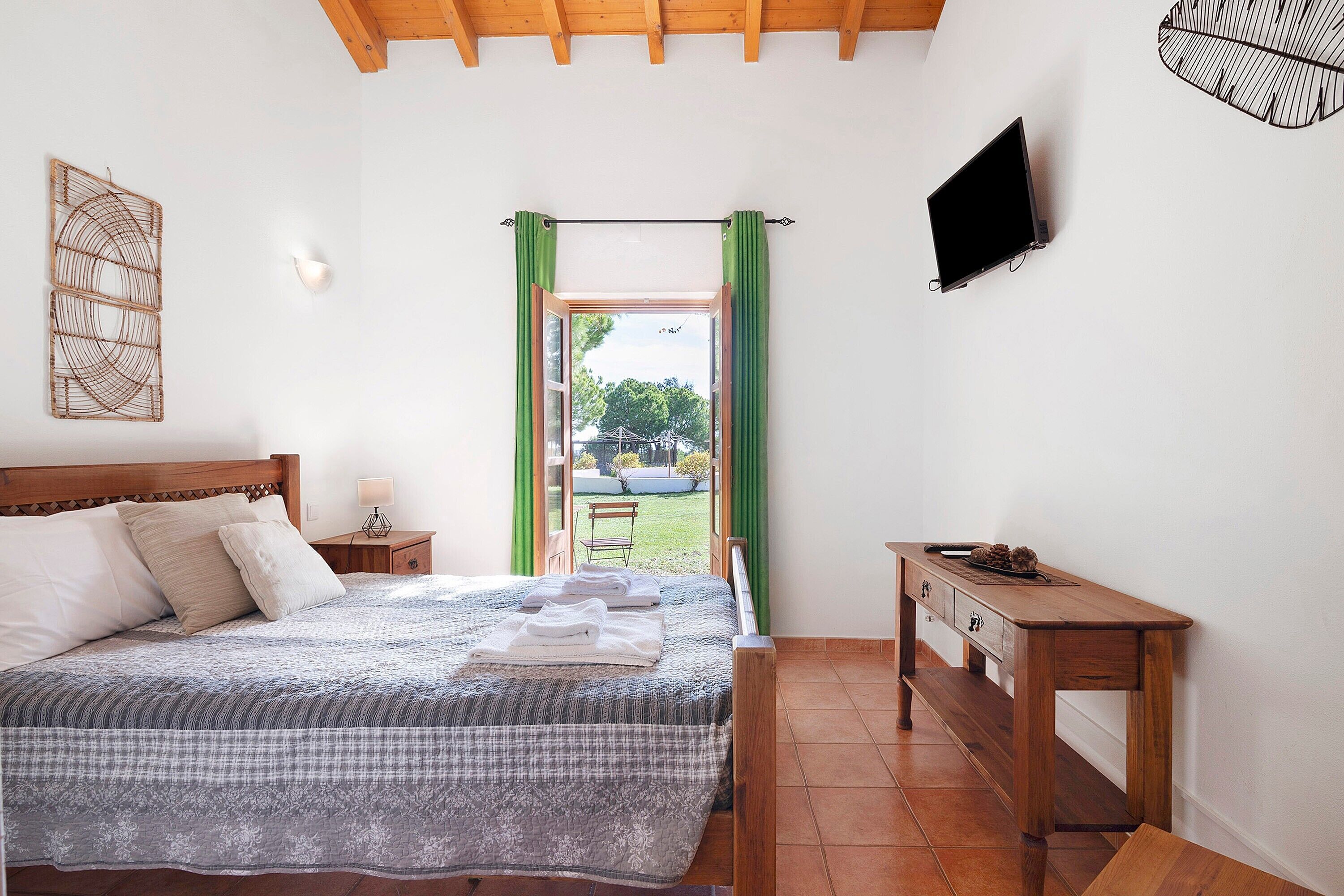 Quarto com cama, mesa de cabeceira de madeira, escrivaninha, televisão e vista pela janela com cortinas.