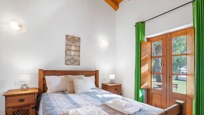 A bedroom with a wooden bed, two bedside tables, a nightstand, a green curtain, and a window with a view of trees. - Casa de Campo Vale do Asno-Room 'Quarto 2' with Shared Pool, Wi-Fi and Air Conditioning (Castro Marim)