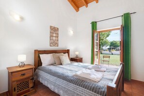 A bedroom with a wooden bed, a nightstand, a wall-mounted lamp, and a view of a garden through an open door.