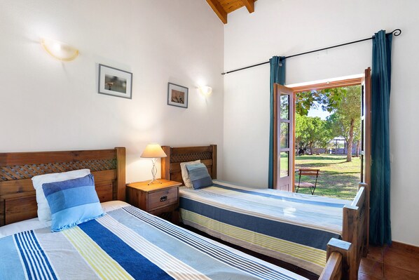 A bedroom with two beds, a wooden headboard, a nightstand, a lamp, and a view of the outdoors. - Casa de Campo Vale do Asno-Room 'Quarto 4' with Shared Pool, Wi-Fi and Air Conditioning (Castro Marim)