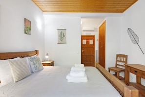 A bedroom with a wooden headboard, a bed with white linens, a nightstand with a lamp, a wooden chair, and a wooden desk.