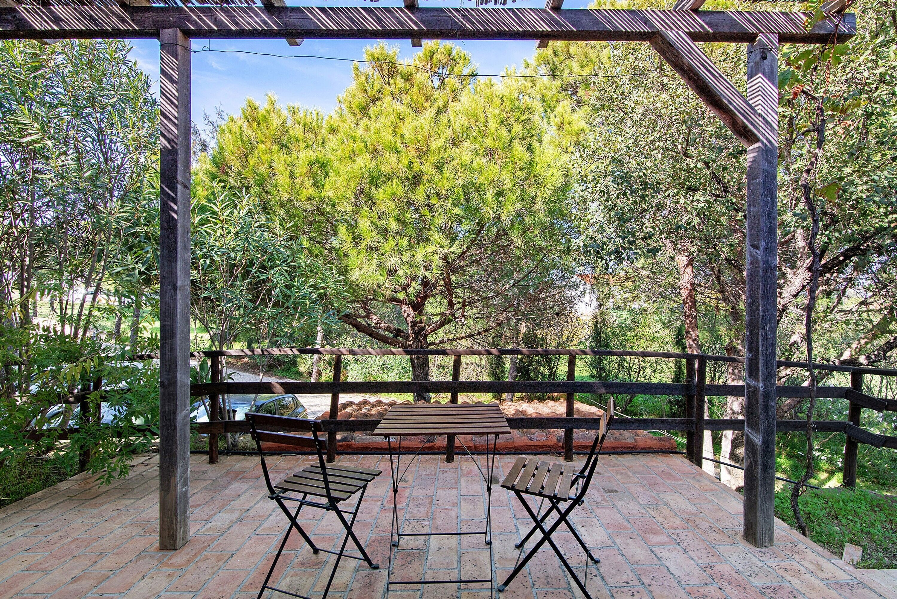 Une terrasse extérieure avec une table en bois et deux chaises, entourée d’une pergola en bois et d’un sol en briques.