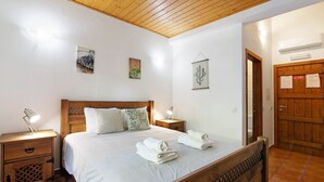A bedroom with a wooden bed, white bedding, a nightstand with a lamp, and a wooden door. - Casa de Campo Vale do Asno-Room 'Quarto 5' with Shared Pool, Wi-Fi and Air Conditioning (Castro Marim)