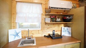A wooden cabin kitchen with a window, sink, and a white air conditioner on the wall. - Tiny Home Retreat with Bunnies, Chickens & a Goat near Gulf Coast Beach Navarre (Navarre)