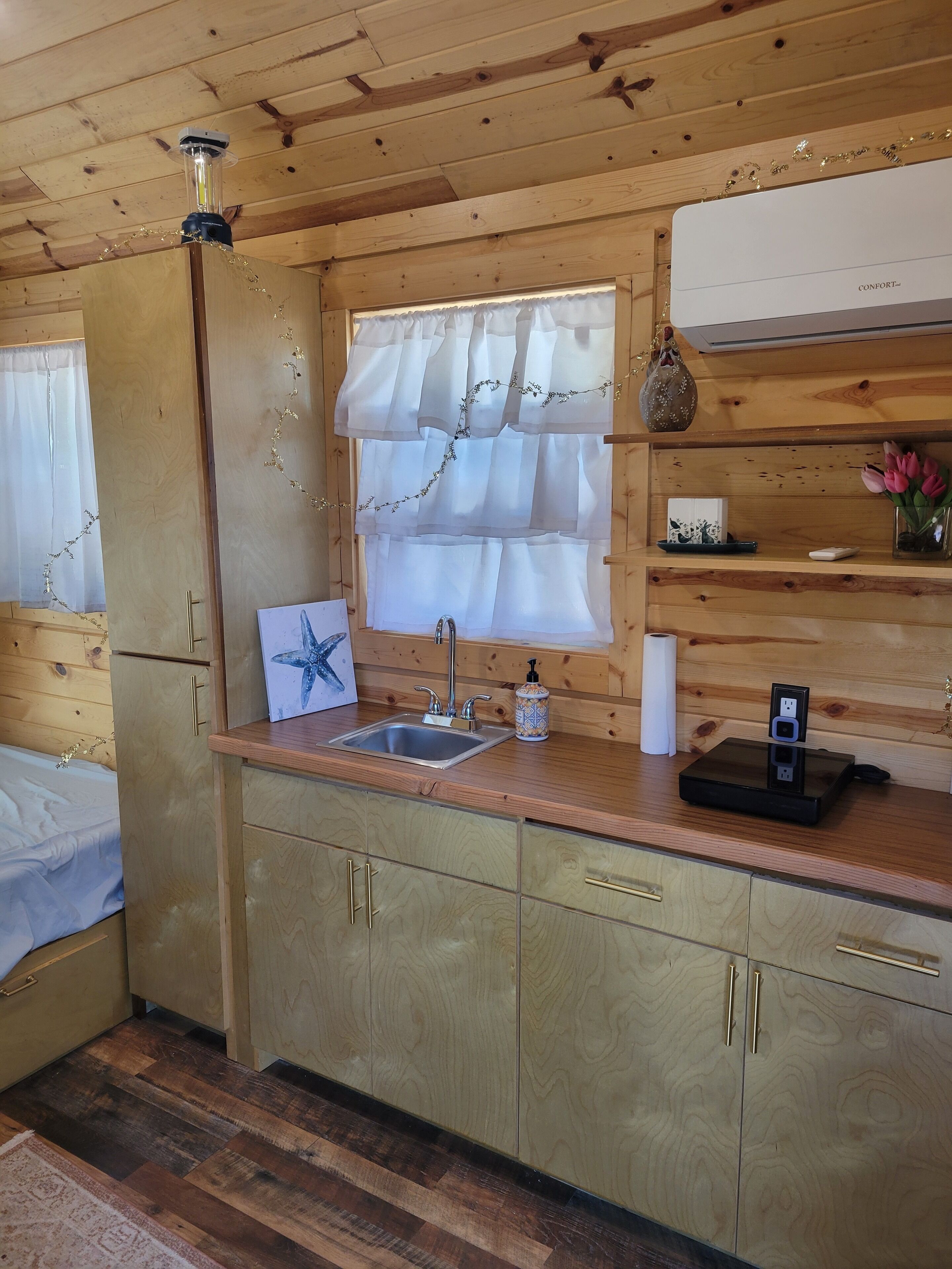 A compact kitchen with a sink, a window with sheer curtains, a small shelf with decorative items, and a wooden ceiling.