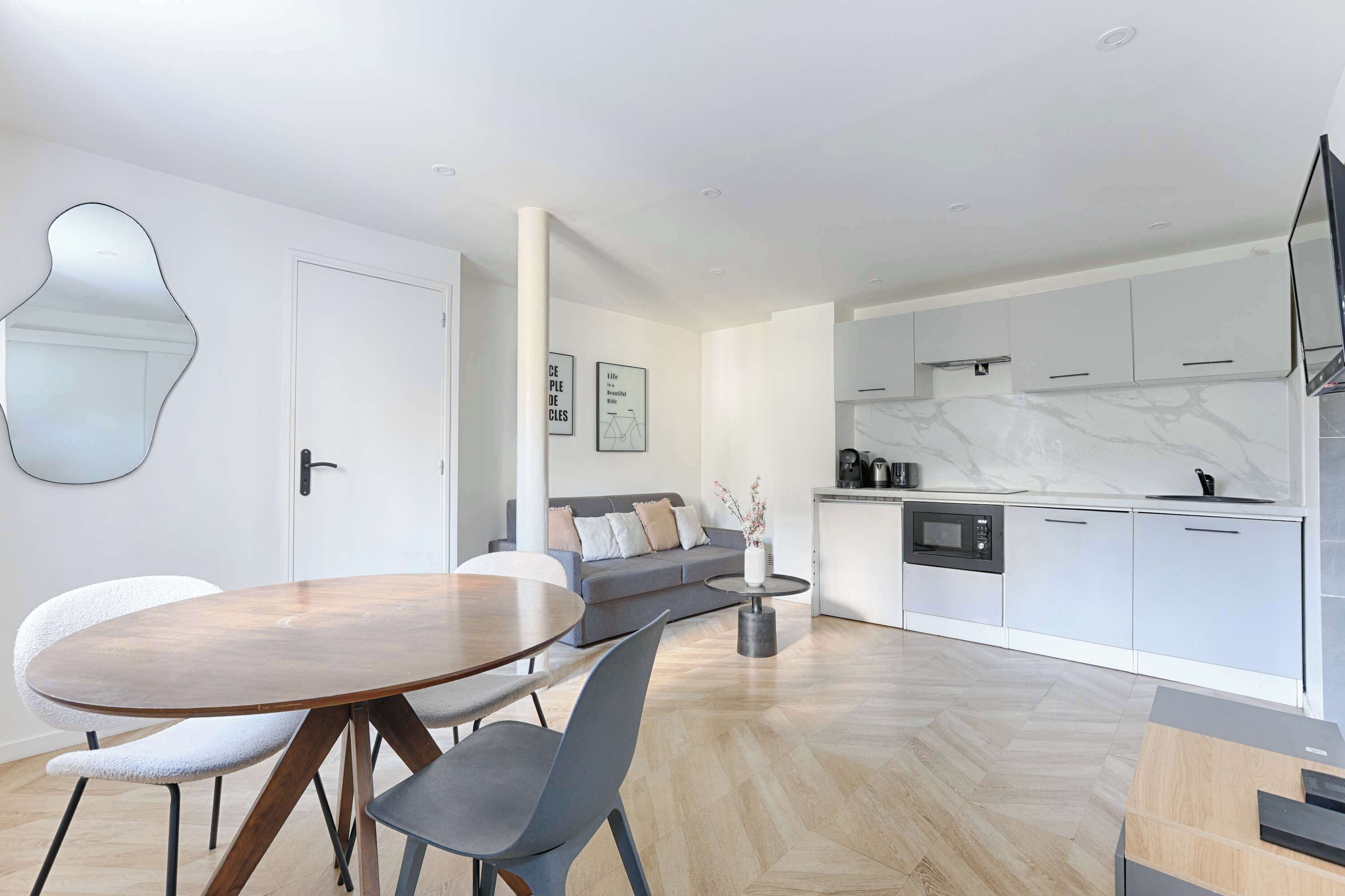 Un espacio de estar moderno con comedor, sofá, cocina con armarios blancos y un espejo grande en la pared.