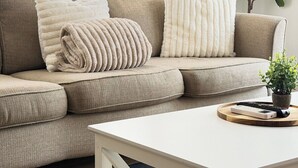 A beige sectional sofa with two pillows, a white coffee table with a remote control, and a potted plant. - Stylish EaDo Apt | Restaurants, Stadiums, Venues (Houston)