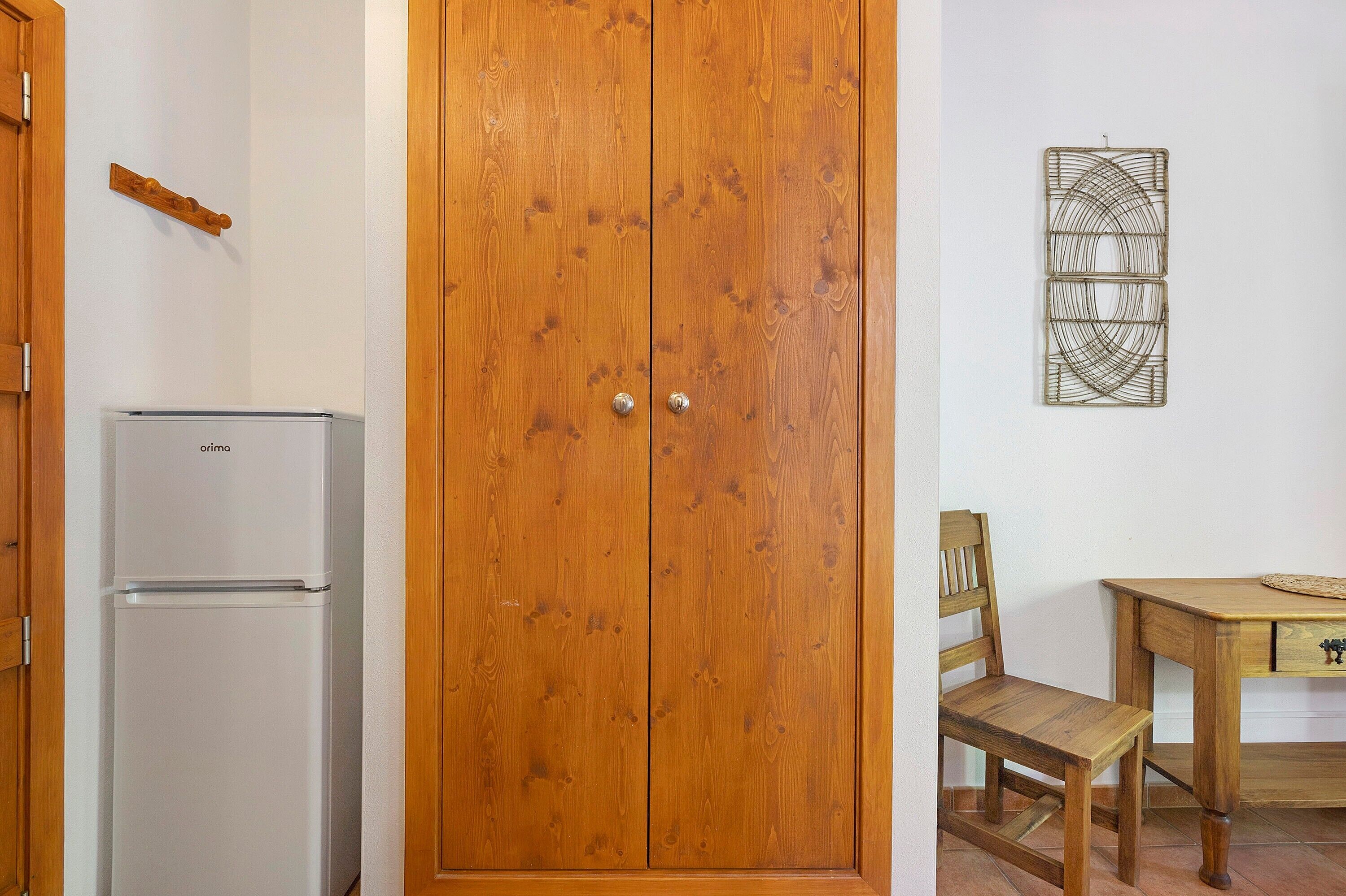 Een houten garderobe, een witte koelkast, een houten stoel en een tafel met een geweven placemat.