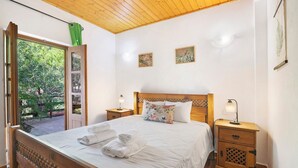 Un dormitorio con cabecera de madera, cama con ropa de cama blanca, dos mesitas de noche con lámparas y vistas a la vegetación a través de una puerta abierta.