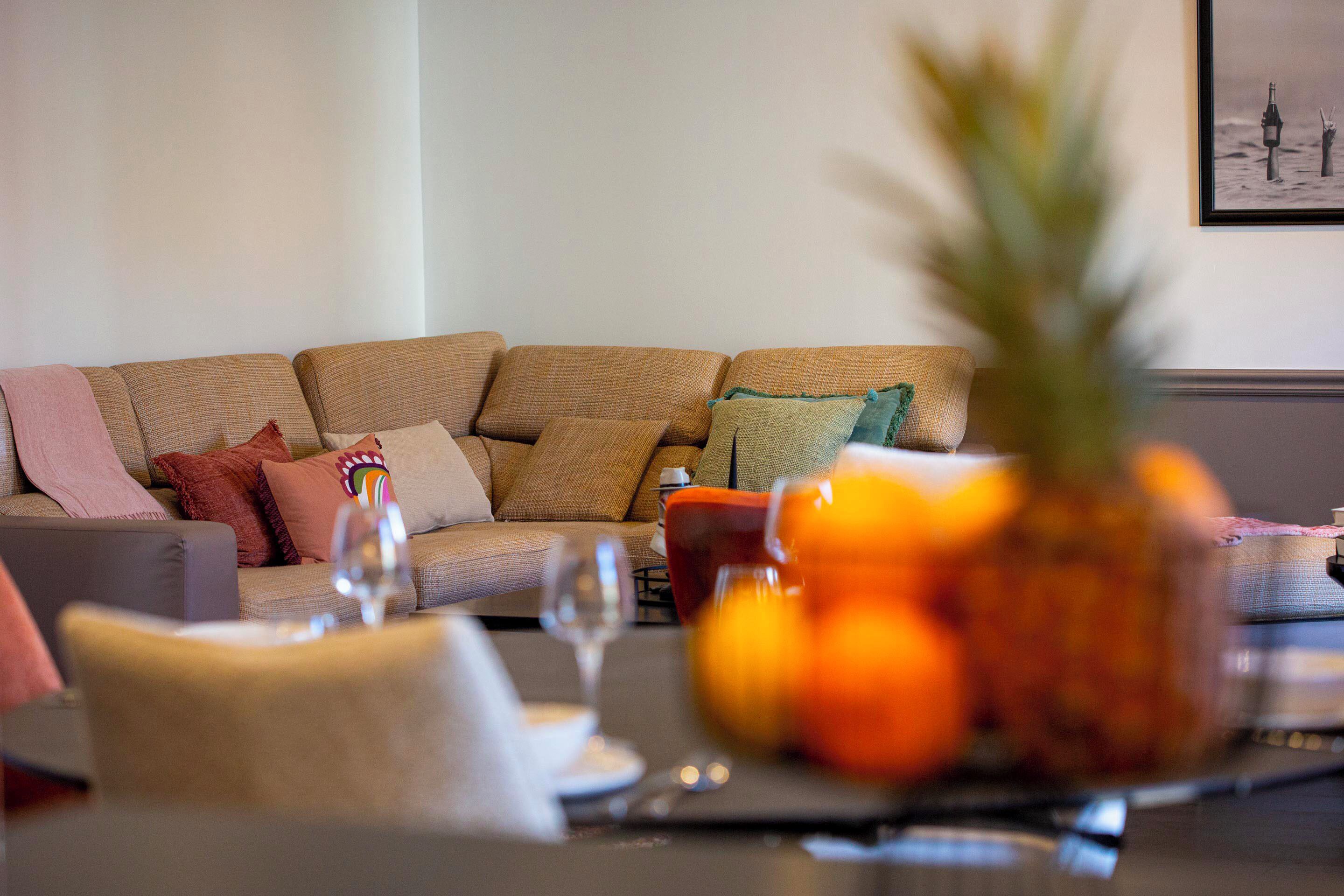 A living room with a sofa, a coffee table with a pineapple and oranges, and a framed picture on the wall.