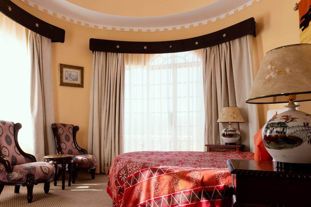 Une chambre d’hôtel avec un grand lit, deux fauteuils, une petite table, une lampe et une fenêtre avec des rideaux.