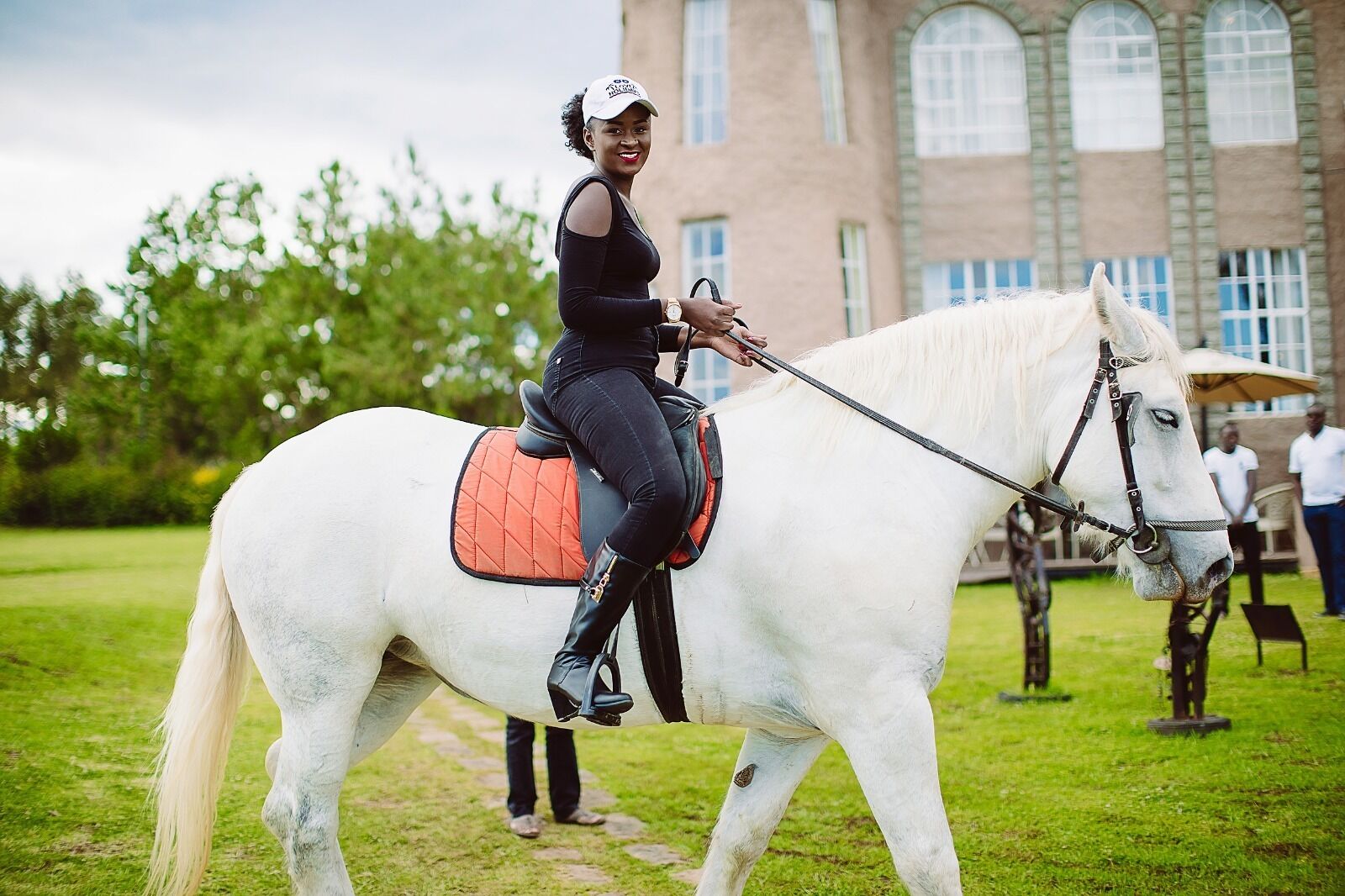 A person riding a white horse with a saddle and bridle, on a grassy path with a building in the background.
