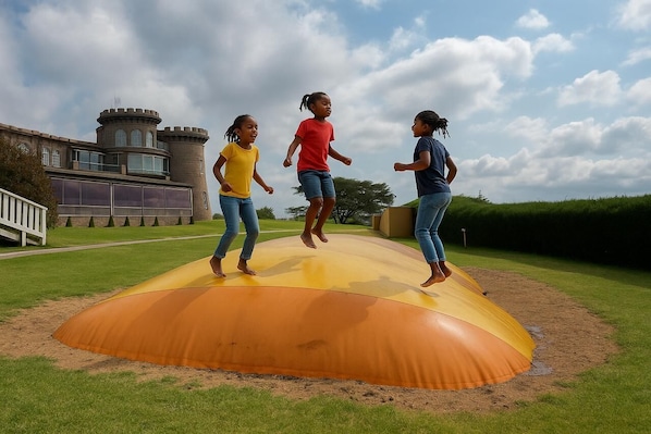 Three children jumping on a large, curved, yellow and orange structure in a grassy area with a castle-like building in the background. - Tafaria Castle & Center for the Arts (Ndaragwa)