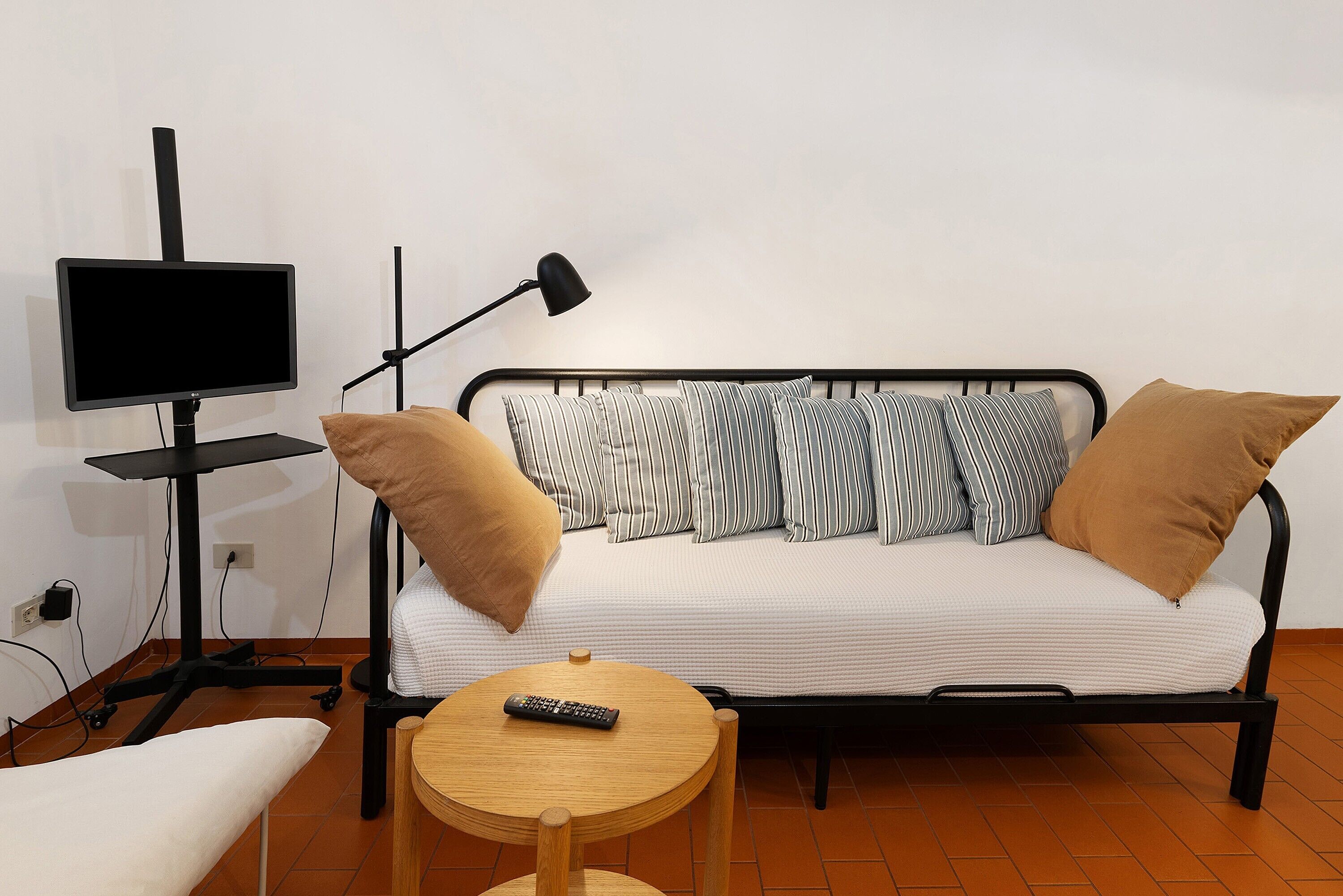 A bed with striped pillows, a remote control on a small table, a standing lamp, and a TV on a stand.