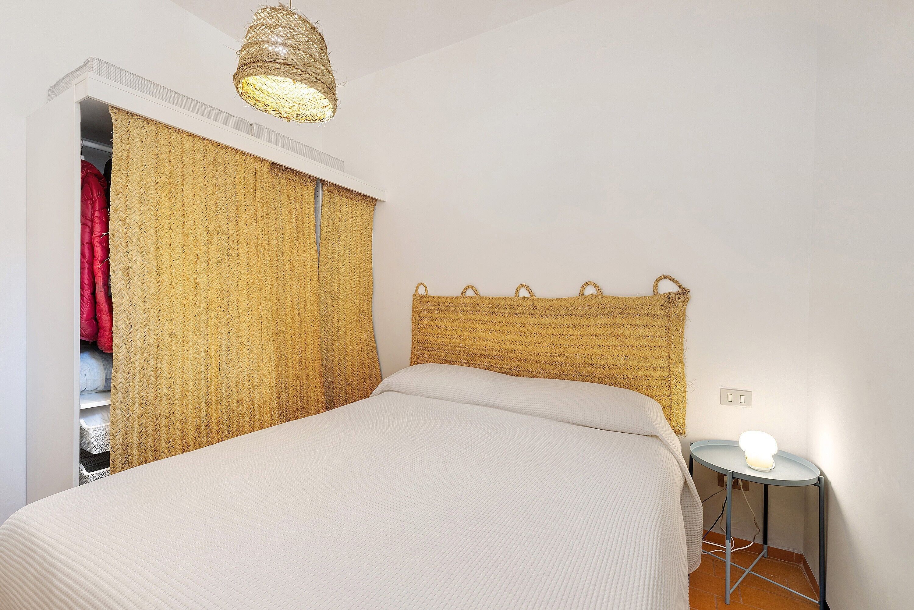 Una camera da letto con un letto, un comodino con una lampada, un armadio pieno di vestiti appesi e una testiera in intreccio.