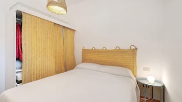 Una camera da letto con un letto, un comodino con una lampada, un armadio pieno di vestiti appesi e una testiera in intreccio.