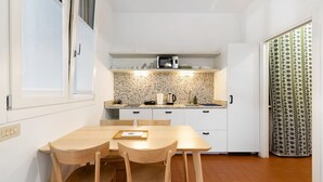 Een compacte keuken met een magnetron, waterkoker en een klein aanrecht. Er is een eettafel met stoelen en een gordijn met een patroon.