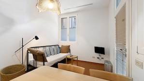 Een kleine kamer met een bed, een eettafel, een televisie en een raam met jaloezieën.