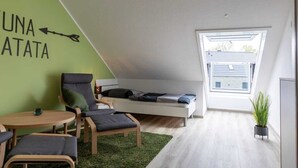 Una habitación moderna y compacta con techo inclinado, una cama, zona de estar con sillas, una mesita pequeña y una planta en maceta.